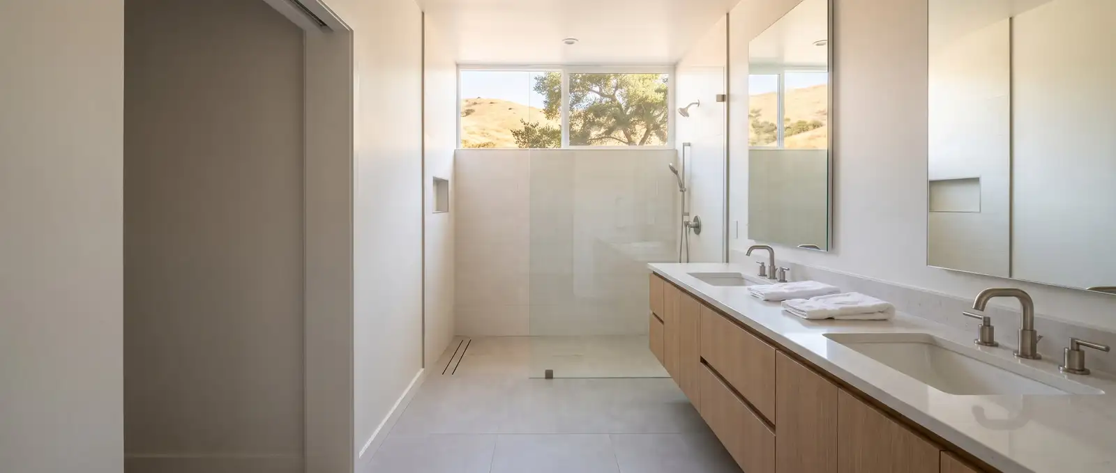 Baño principal con un gabinete doble largo de roble blanco y una regadera de vidrio sin escalón, con cerros dorados por una ventana alta, ilustración generada con IA